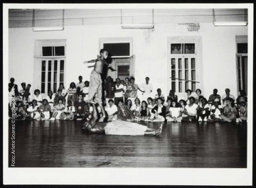 Fotografia de sala com janelas e porta ao fundo. No centro, homem está deitado no chão segurando e levantando nas mãos menino pelos pés. Ao fundo, há pessoas sentadas e em pé assistindo.