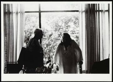 Lina Bo Bardi em pé ao lado de Pierre Verger na Casa de Vidro. Eles olham um para o outro e estão ligeiramente separados e Verger está de perfil, enquanto Bo Bardi está de frente com o rosto voltado para o lado. A fotografia foi tirada contra a luz, o que faz com que seja possível ver somente os contornos dos dois.
