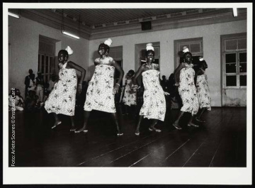 Mulheres vestidas com vestidos brancos com estampas de folhas. Elas usam colares e brincos e ornamentos brancos na cabeça, e parecem ter um tipo de pó branco nos ombros e na região da clavícula. Elas estão posicionadas em círculo, têm as bocas abertas, e estão com as mãos próximas à cintura, no que parece ser um movimento de dança. Ao fundo é possível ver a parede com janelas.