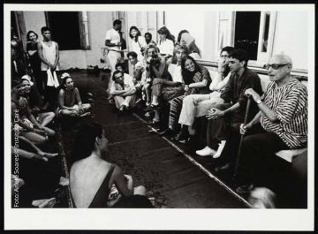 Fotografia de sala que aparenta ser destinada para ensaios com pessoas sentadas e em pé. A maioria está voltada para Pierre Verger, que está sentado segurando bengala. Caetano Veloso está ao lado de Verger.