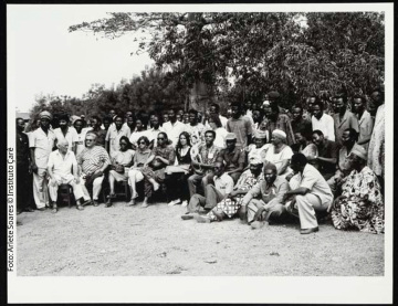 Fileira de pessoas sentadas (Gilberto Gil está entre elas), algumas delas usam óculos escuros. Atrás delas, há multidão de pessoas em pé. Na parte superior da fotografia, é possível ver copas de árvores.